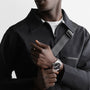 A person wearing a black zip-up jacket and a shoulder strap displays a Longines Conquest wristwatch with automatic movement and a black face on their left wrist.