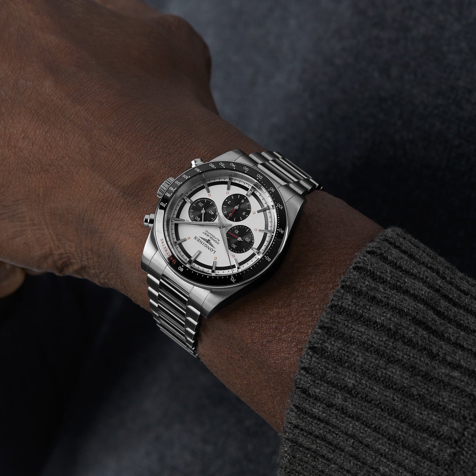 A person wearing the Longines Conquest Chronograph, featuring a white and black dial and self-winding movement, with a ribbed gray sweater sleeve against a dark background.