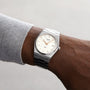 Someone wears a Tissot PRX stainless steel wristwatch with a white dial, silver indices, and date window—its design inspired by the 1970s—all set against a plain background.