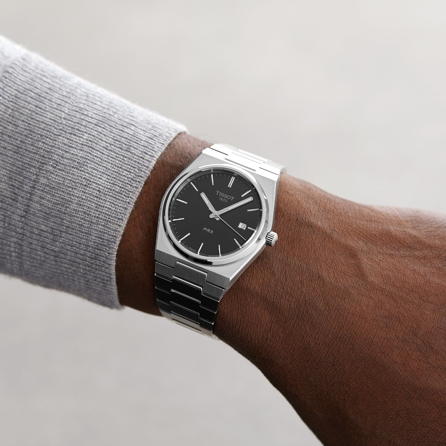 Someone wears a Tissot PRX with Swiss quartz movement, silver stainless steel case, black face, and date display, paired with a light gray long-sleeve shirt against a neutral background.