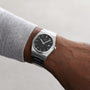 Someone wears a Tissot PRX with Swiss quartz movement, silver stainless steel case, black face, and date display, paired with a light gray long-sleeve shirt against a neutral background.