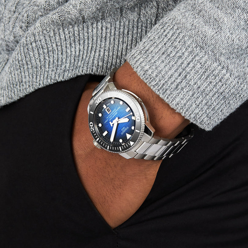 A person wearing a Tissot Seastar 2000 Professional Powermatic 80 watch with a blue face and black bezel, paired with a grey sweater and black pants.