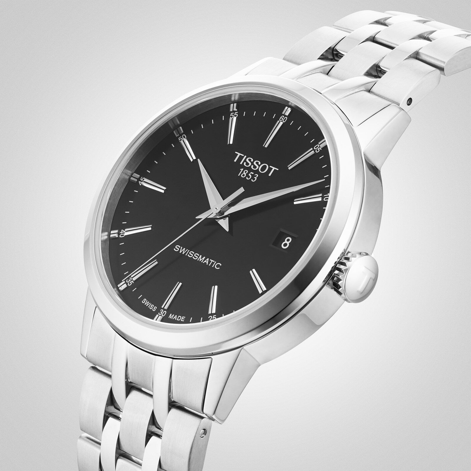 The Tissot Classic Dream Swissmatic is a silver wristwatch with a black dial, date window, and metal link band, featuring Swissmatic automatic movement and displayed at an angle on a light gray background.