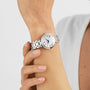A person wearing a Tissot Bellissima Automatic silver wristwatch with a white face and Roman numerals holds their wrist with their other hand.