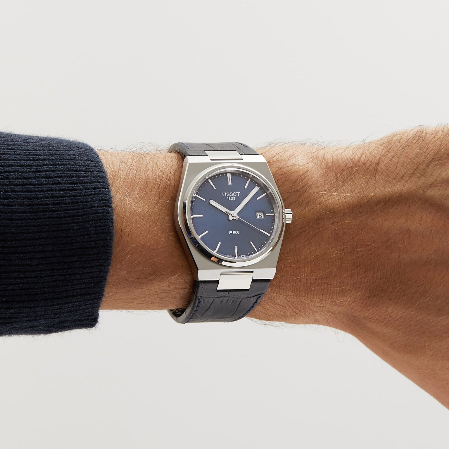 A Tissot PRX watch, inspired by the 1970s, with a blue dial and silver case is worn on the left wrist, paired with a dark textured strap.
