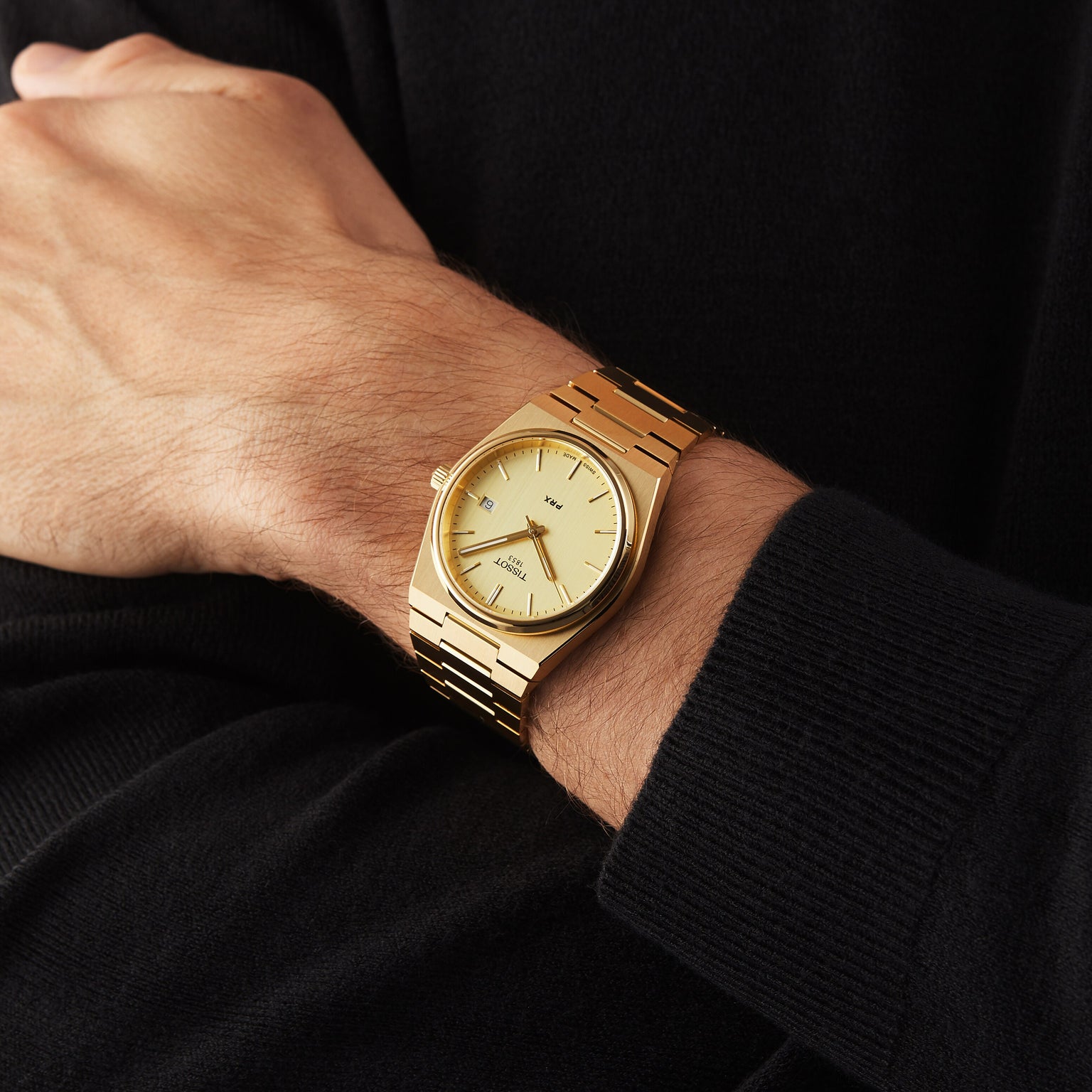 A person wears a Tissot PRX gold wristwatch with a matching gold band and Swiss quartz movement, paired with a black long-sleeve shirt.