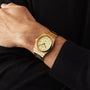 A person wears a Tissot PRX gold wristwatch with a matching gold band and Swiss quartz movement, paired with a black long-sleeve shirt.
