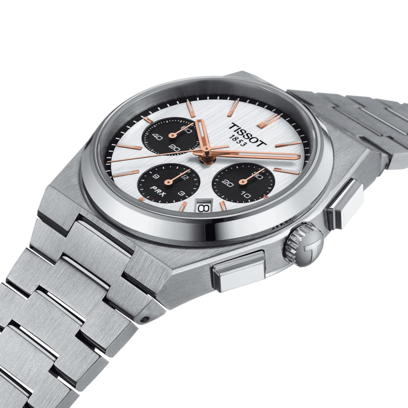 The Tissot PRX Automatic Chronograph features a stainless steel case, silver dial with three black subdials, rose gold hands, and a metal link bracelet, inspired by 70s design for a timeless look.