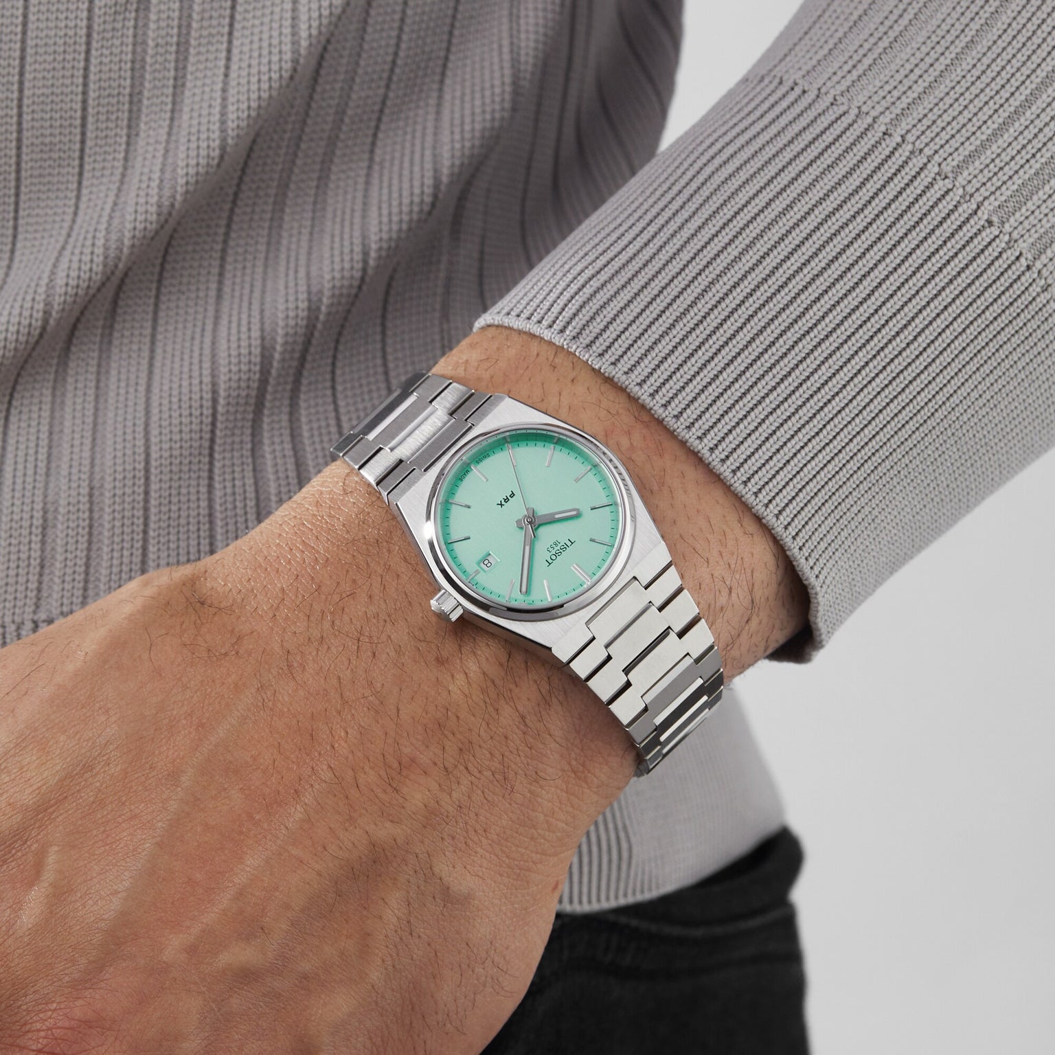 A person wearing the Tissot PRX, a silver metal retro watch with a mint green dial, and a ribbed, light gray long-sleeve shirt.