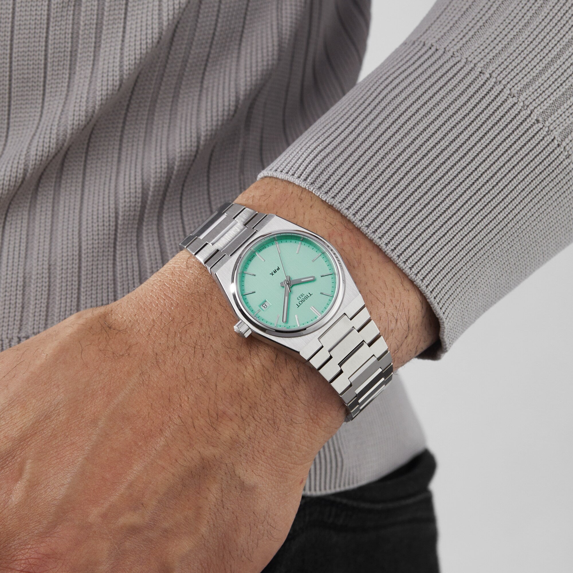 A person wearing the Tissot PRX, a silver metal retro watch with a mint green dial, and a ribbed, light gray long-sleeve shirt.