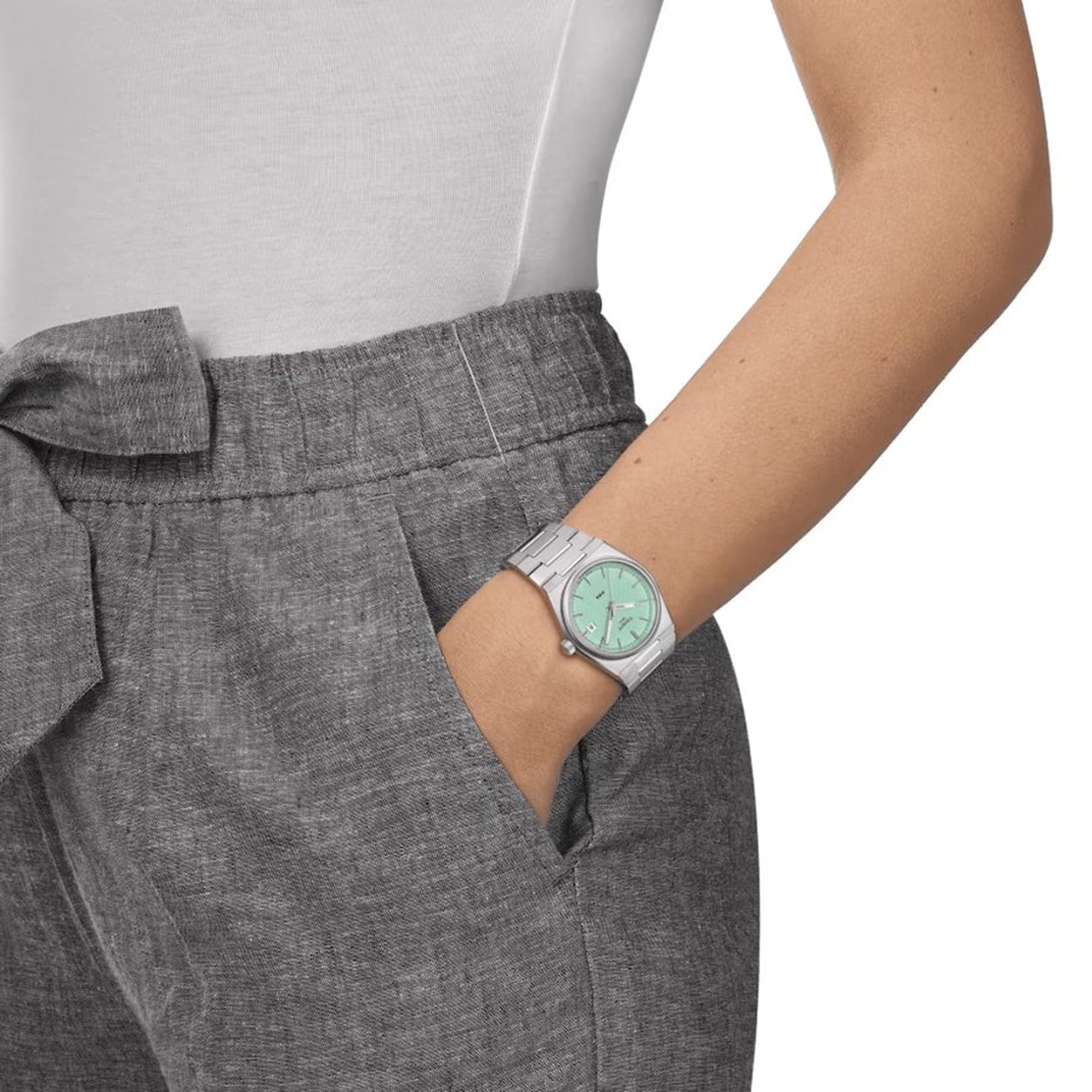 A person in grey pants and a white shirt has their hand in their pocket, showing off the Tissot PRX watch with a mint green dial.