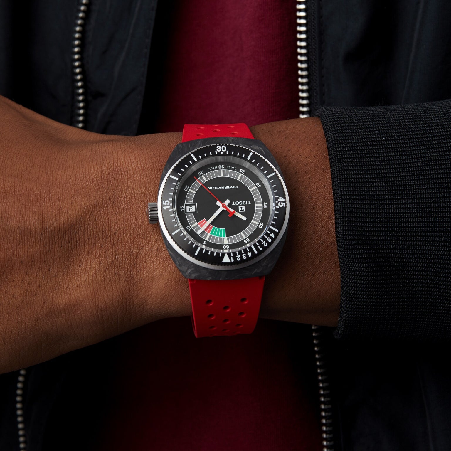 A person in a black jacket showcases a Tissot Sideral S Powermatic 80 watch with a round dial and red strap on their wrist.