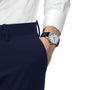 A person wearing a white shirt, navy trousers, and a Tissot Le Locle Powermatic 80 20th Anniversary wristwatch with automatic movement, blue strap, and white face has their hand in their pocket.
