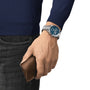 Someone dressed in a navy sweater and dark jeans holds a brown leather wallet in their left hand while wearing a Tissot PR516 Chronograph with a blue dial.