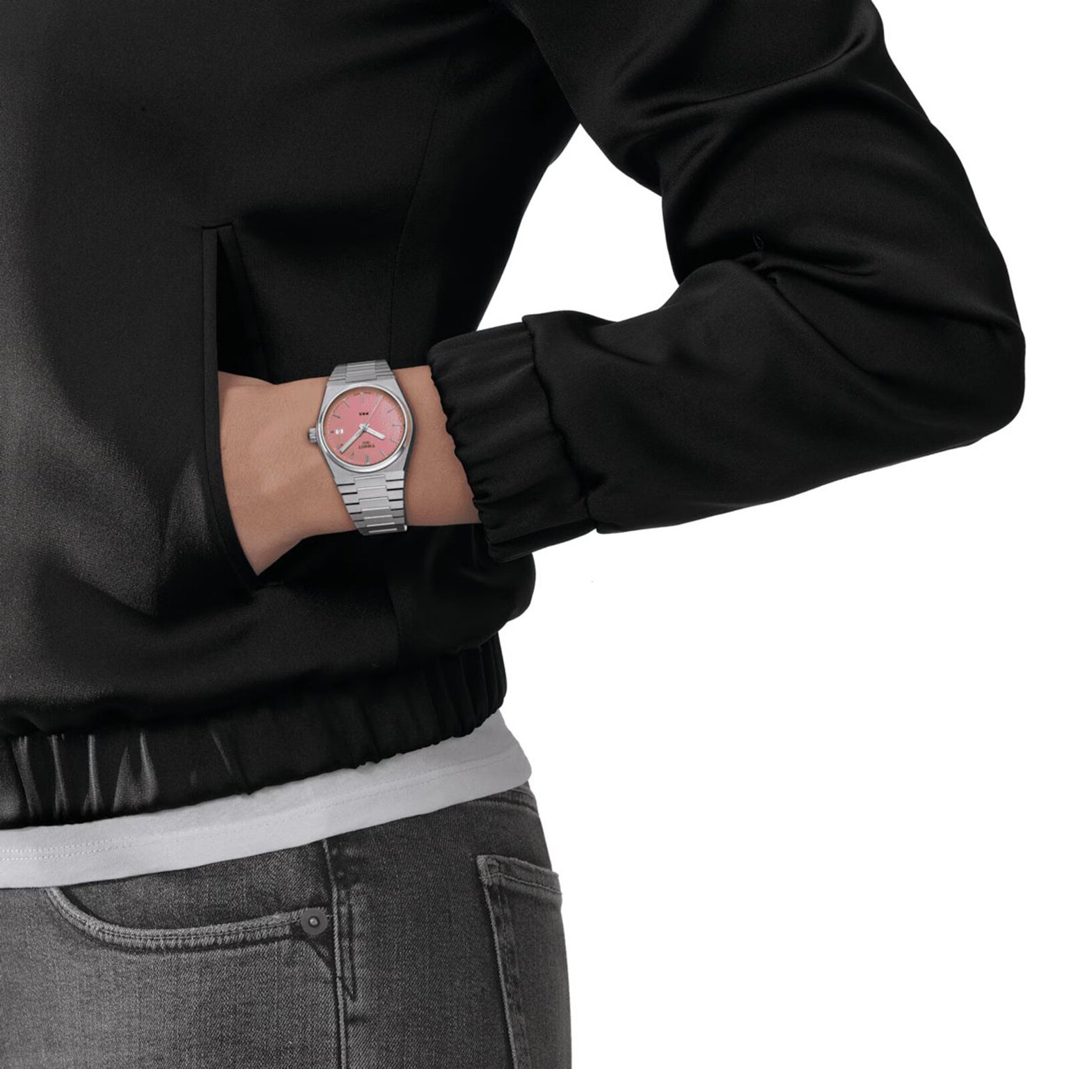 A person in a black jacket and jeans with their left hand in their pocket showcases the Tissot PRX, a retro watch featuring a pink face and stainless steel bracelet.