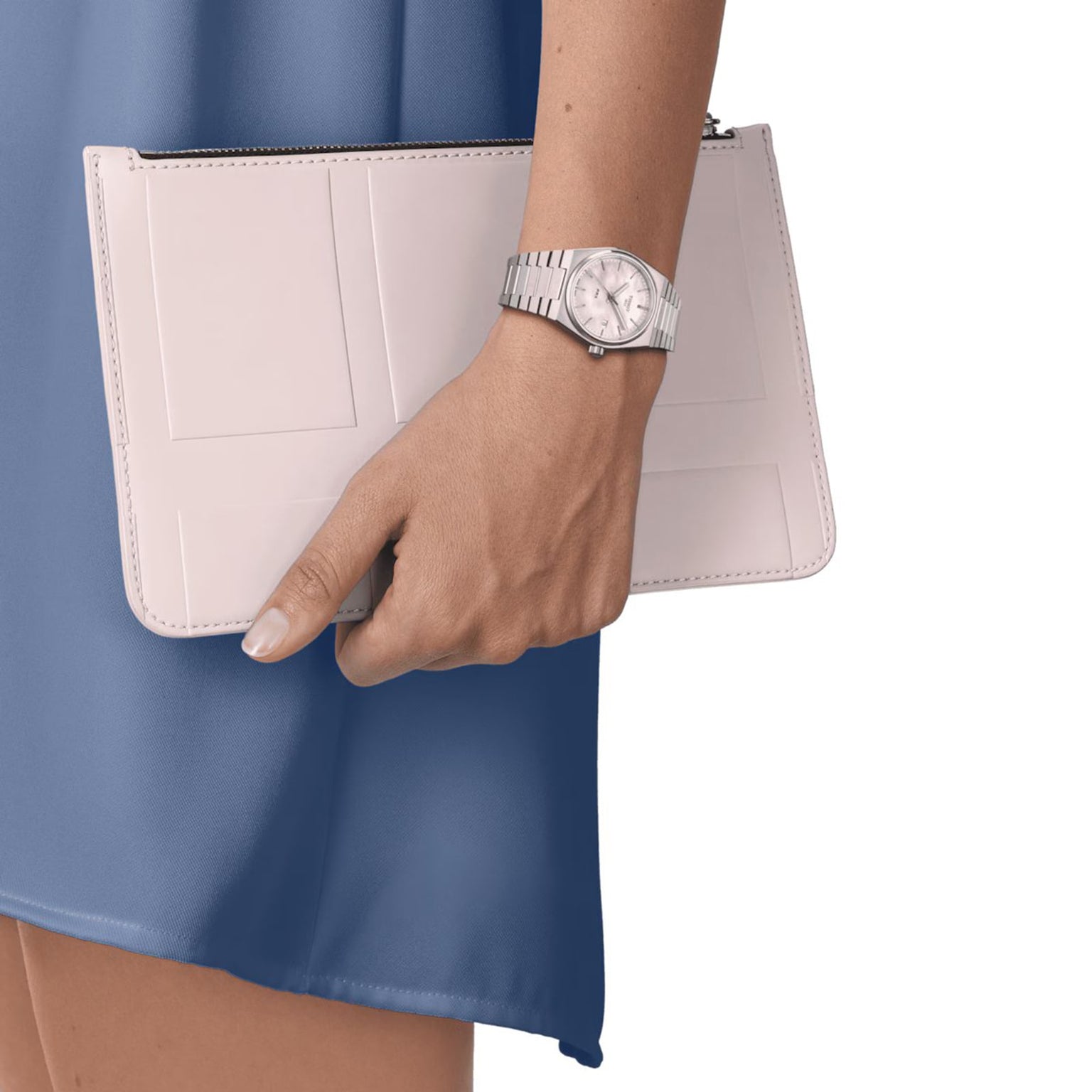 A person in a blue dress holds a beige clutch and wears the Tissot PRX silver wristwatch on their left hand.