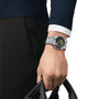 A person in a dark blue sweater and black pants holds a black handle, displaying a sleek Tissot PRX Automatic Chronograph watch with a black dial on their left wrist.