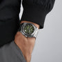 Wearing a Tissot PRX Automatic Chronograph with a silver band and green dial, their hand is partly in their pocket, dressed in a black jacket and gray pants.