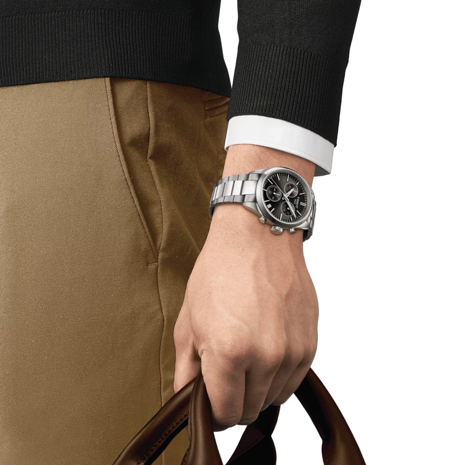 A person in a black long-sleeve shirt and tan pants holds a brown leather bag; on their left wrist, they wear the Tissot PR 100 Chronograph, a silver quartz watch with Super-LumiNova® coating for a sporty touch.