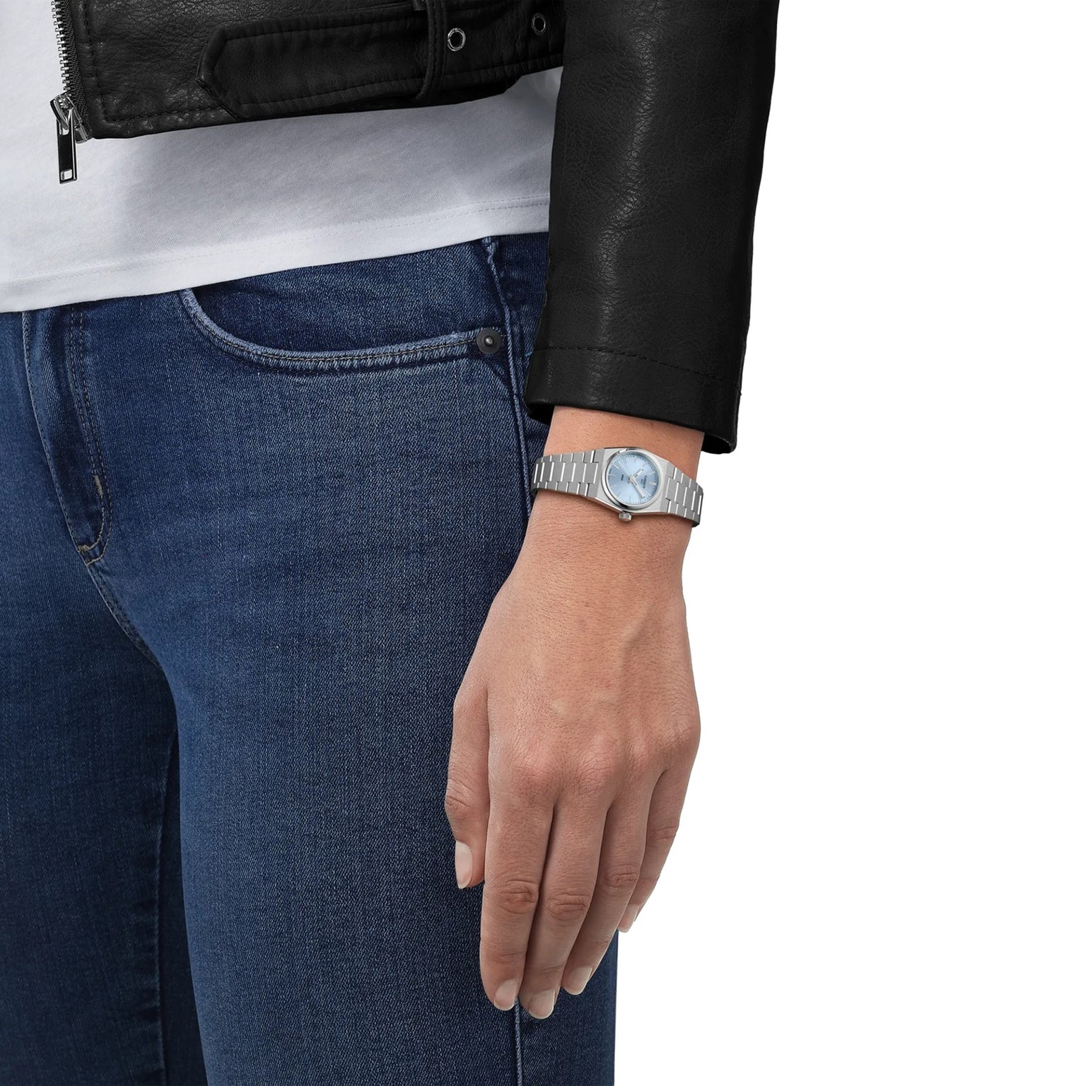 A person stands with hand by their side, wearing blue jeans, a black leather jacket, and a Tissot PRX silver wristwatch.