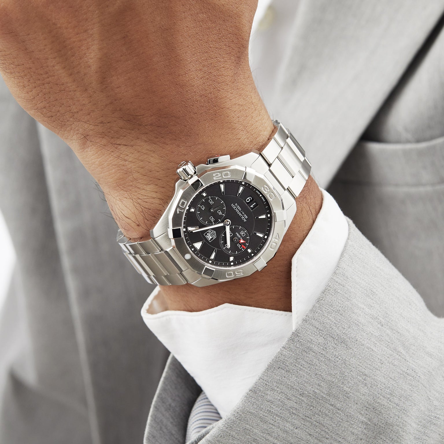 A person in a light gray suit and white shirt wears a TAG Heuer Aquaracer sports watch with water resistance, featuring a black dial and silver bezel.