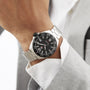 A person in a light gray suit wears a TAG Heuer Formula 1 Date wristwatch with a black dial, white numerals, quartz movement, date display, and stainless steel case on their left wrist.