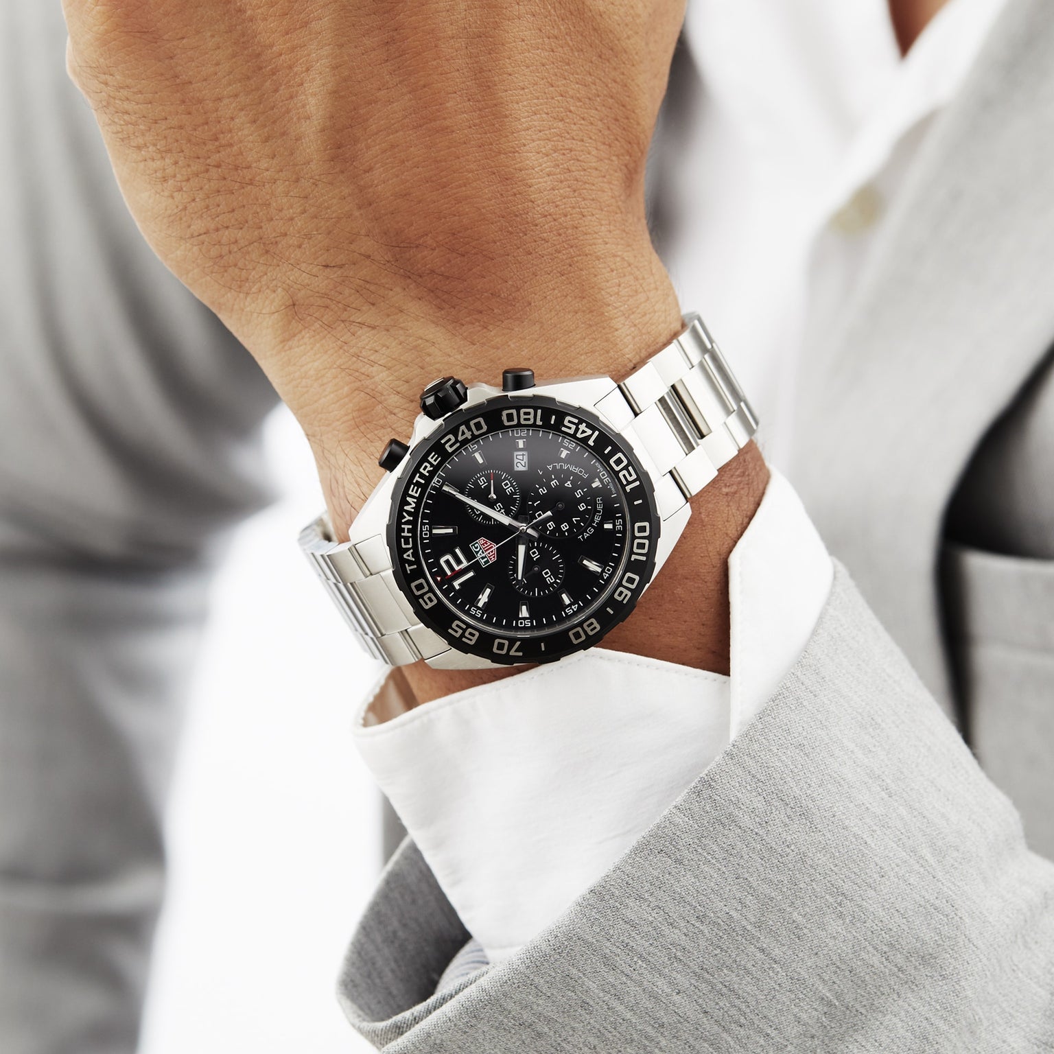 A person in a light gray suit wears a TAG Heuer Formula 1 Chronograph wristwatch with a black face and tachymeter bezel.