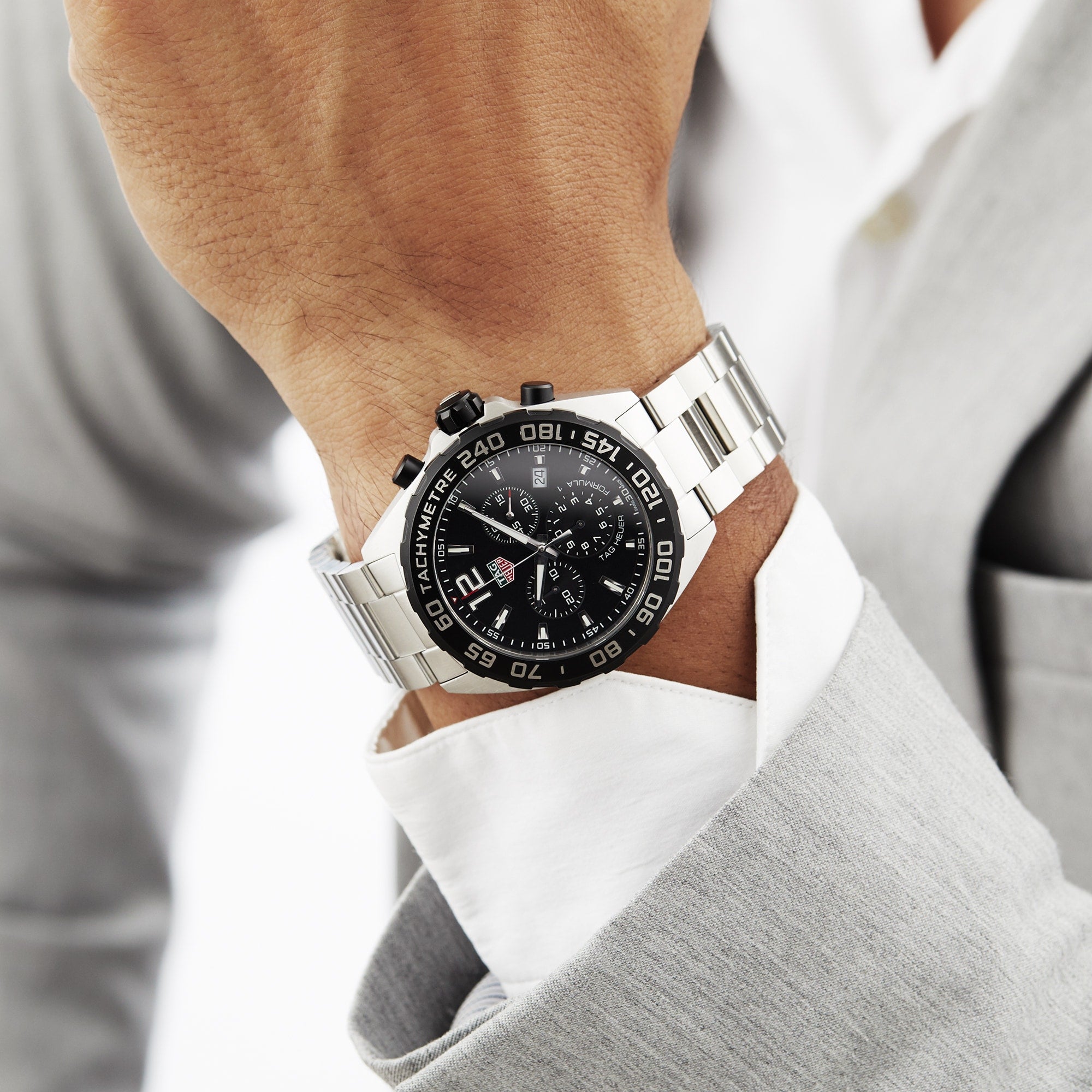 A person in a light gray suit wears a TAG Heuer Formula 1 Chronograph wristwatch with a black face and tachymeter bezel.