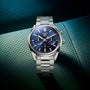 The TAG Heuer Carrera Chronograph, featuring a blue dial, chronograph subdials, and date display, is set on a textured teal surface, highlighting its motor-racing heritage.