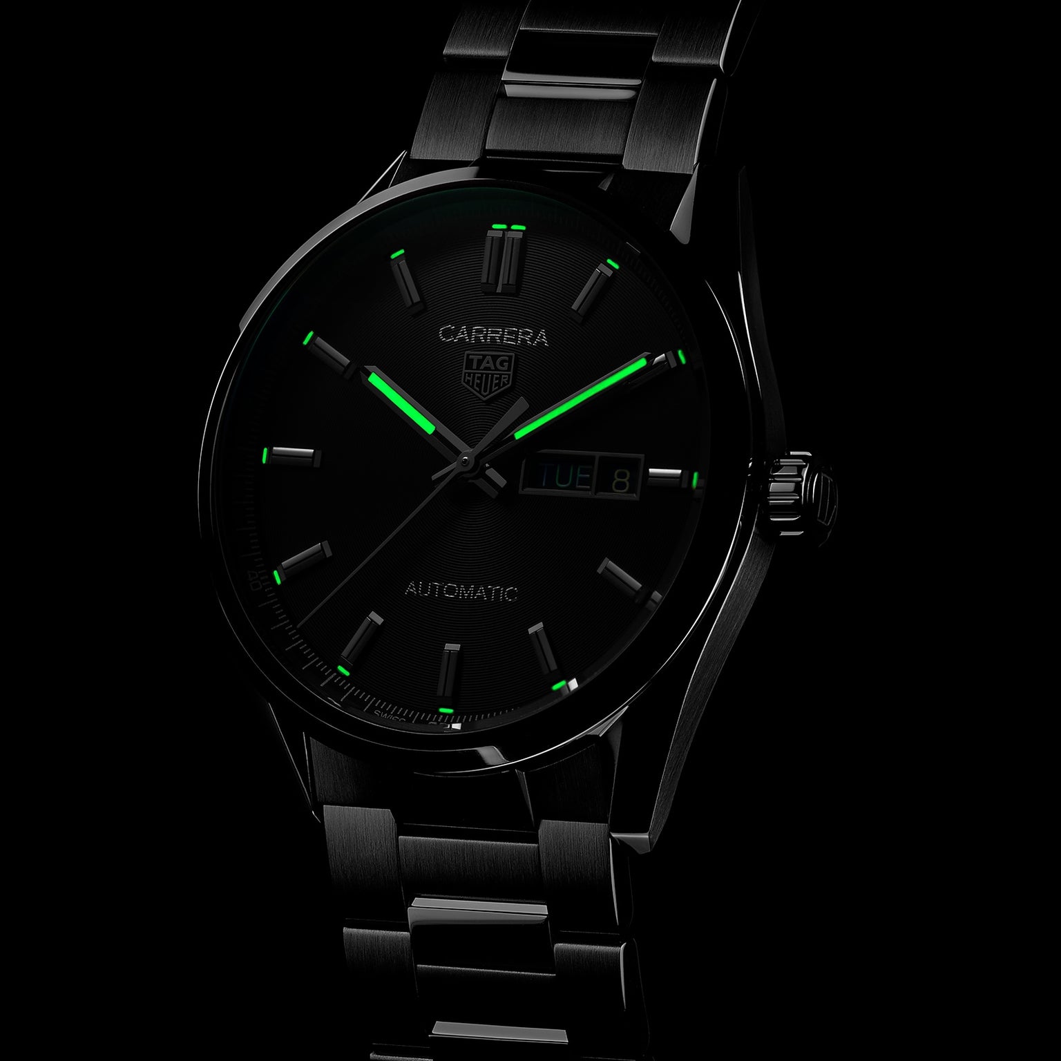 The TAG Heuer Carrera Day-Date wristwatch features a dark stainless steel case, black azurage dial, green luminescent hour markers and hands, day-date display, all partially illuminated against a black background.