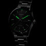 The TAG Heuer Carrera Day-Date wristwatch features a dark stainless steel case, black azurage dial, green luminescent hour markers and hands, day-date display, all partially illuminated against a black background.