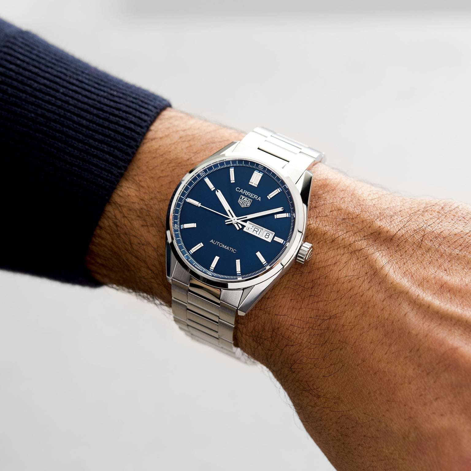 A TAG Heuer Carrera Day-Date stainless steel watch with a blue sunray dial, silver hour markers, and metal band is worn on a left wrist, featuring automatic movement and displaying the day and date.