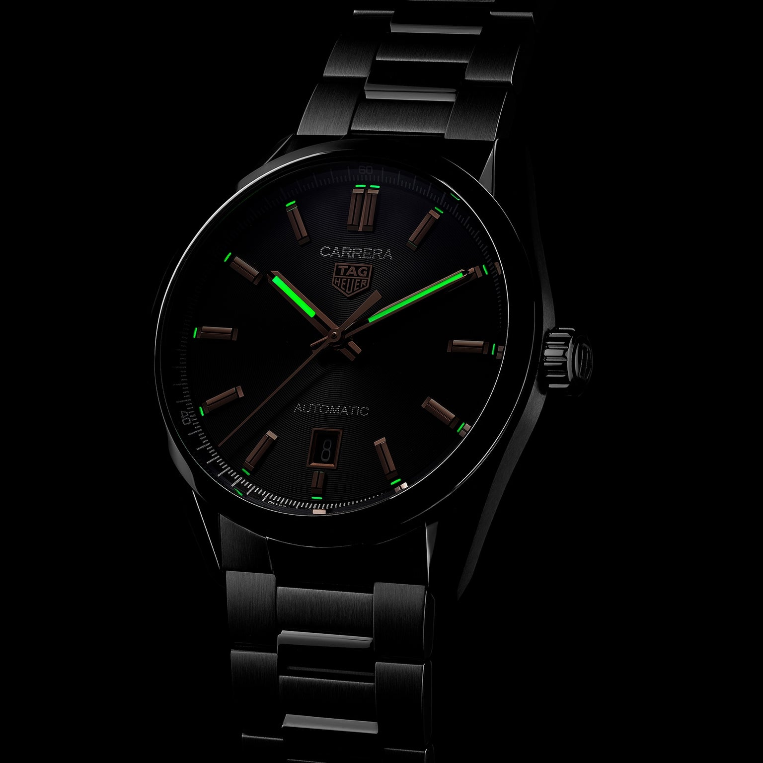 The TAG Heuer Carrera Date watch features a black dial, steel case, and metal band. In low light, its green luminescent hour markers and hands stand out. Powered by the precision Calibre 5 movement.