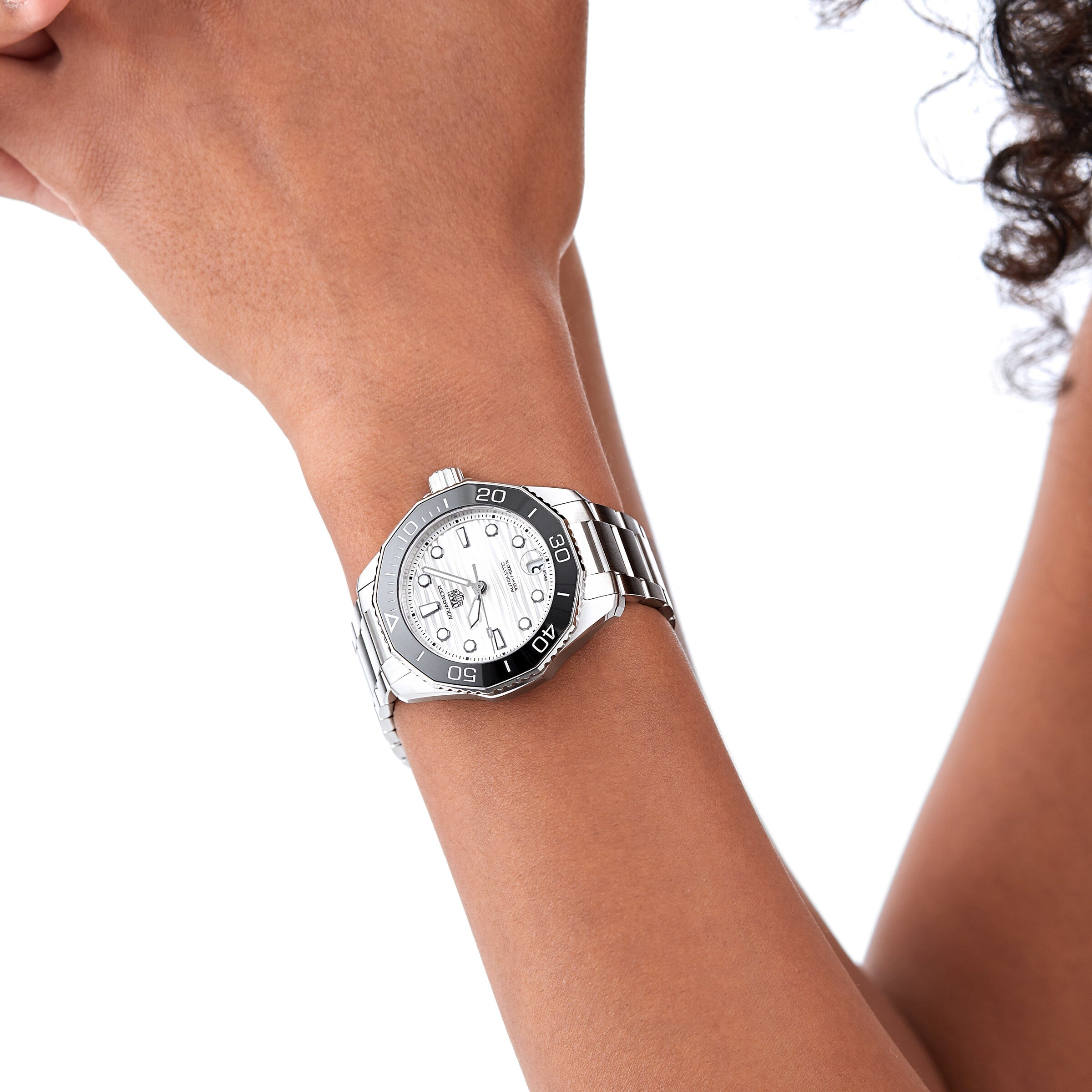 A person with medium brown skin wears the TAG Heuer Aquaracer Professional 300 Date, a sporty silver wristwatch with a white face and black bezel, displayed against a white background.