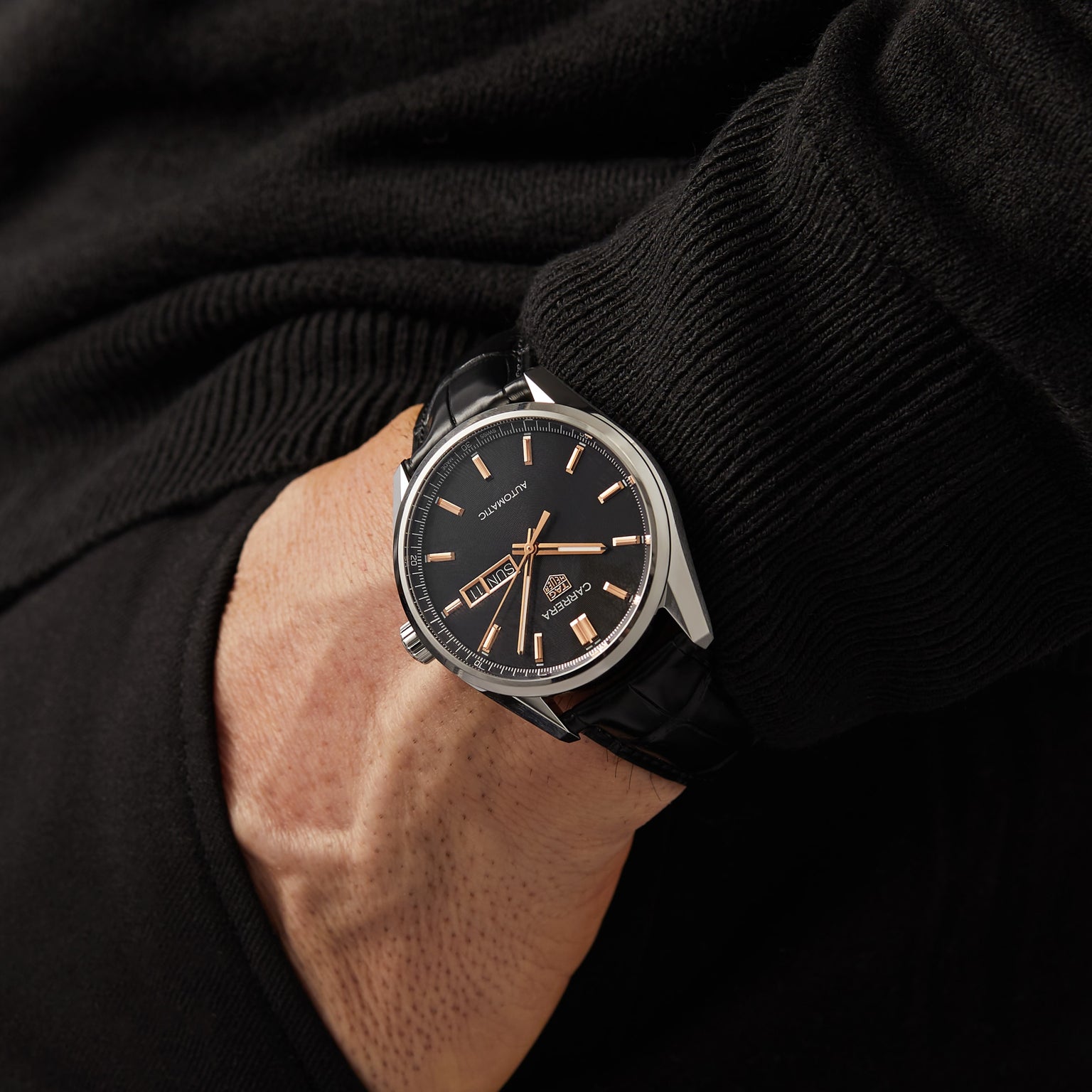 Wearing the TAG Heuer Carrera Day-Date with a black leather band and silver case, paired with a black long-sleeve shirt and hand in pocket, exudes a refined luxury aesthetic.