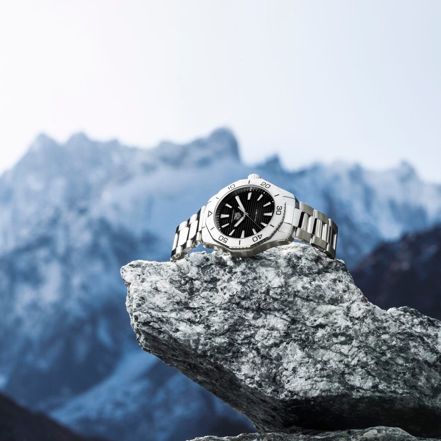 The TAG Heuer Aquaracer Professional 200, a silver sports watch with black dial and stainless steel case, is displayed on a gray rock against a backdrop of snow-capped mountains.