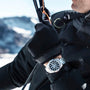 A person in a black jacket and gloves holds a strap outdoors, showcasing the TAG Heuer Aquaracer Professional 200 watch with a stainless steel case on their wrist against snowy mountains.