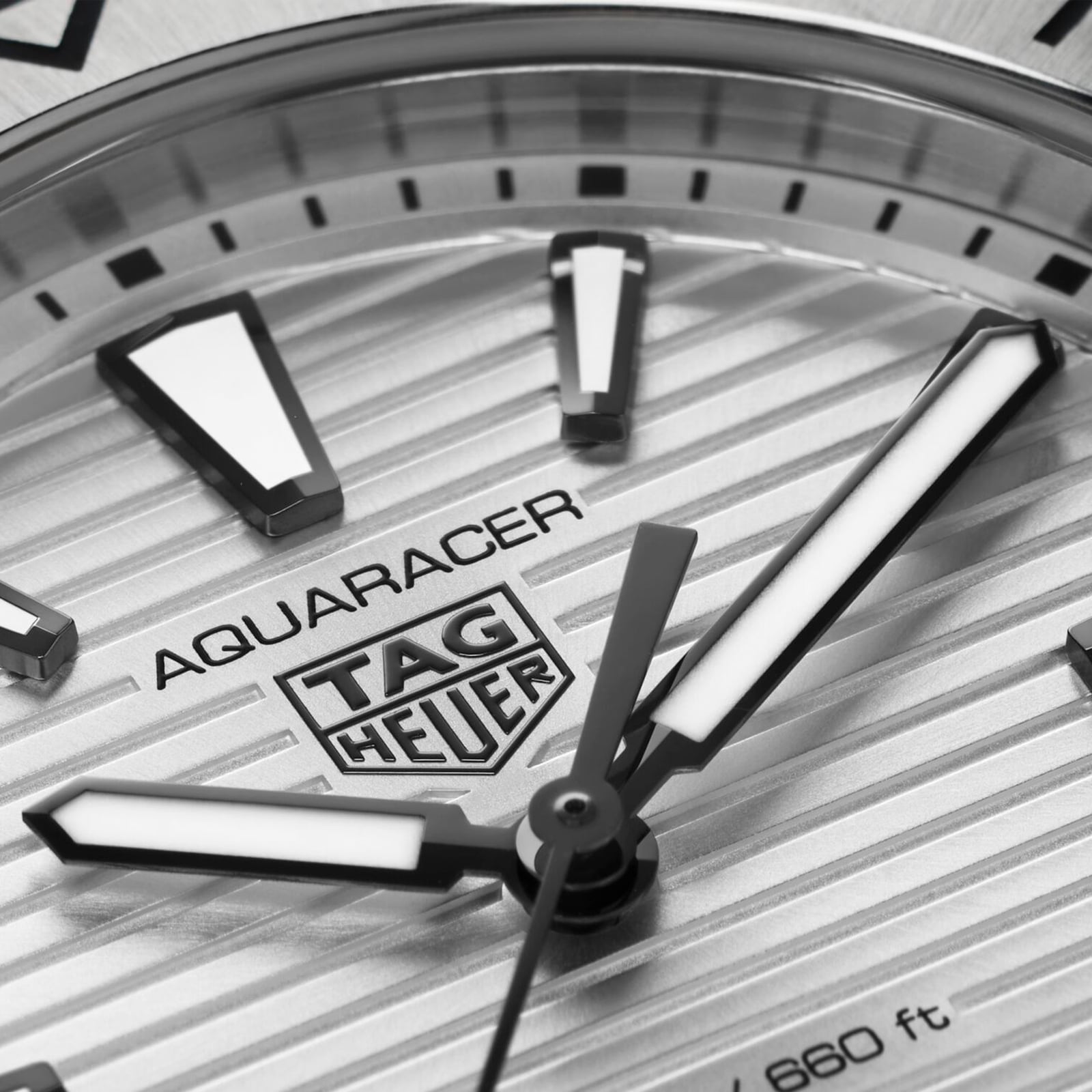 Close-up of a TAG Heuer Aquaracer Professional 200 watch face featuring a striped texture, bold luminous hour markers, stainless steel case, and partially visible hour and minute hands.