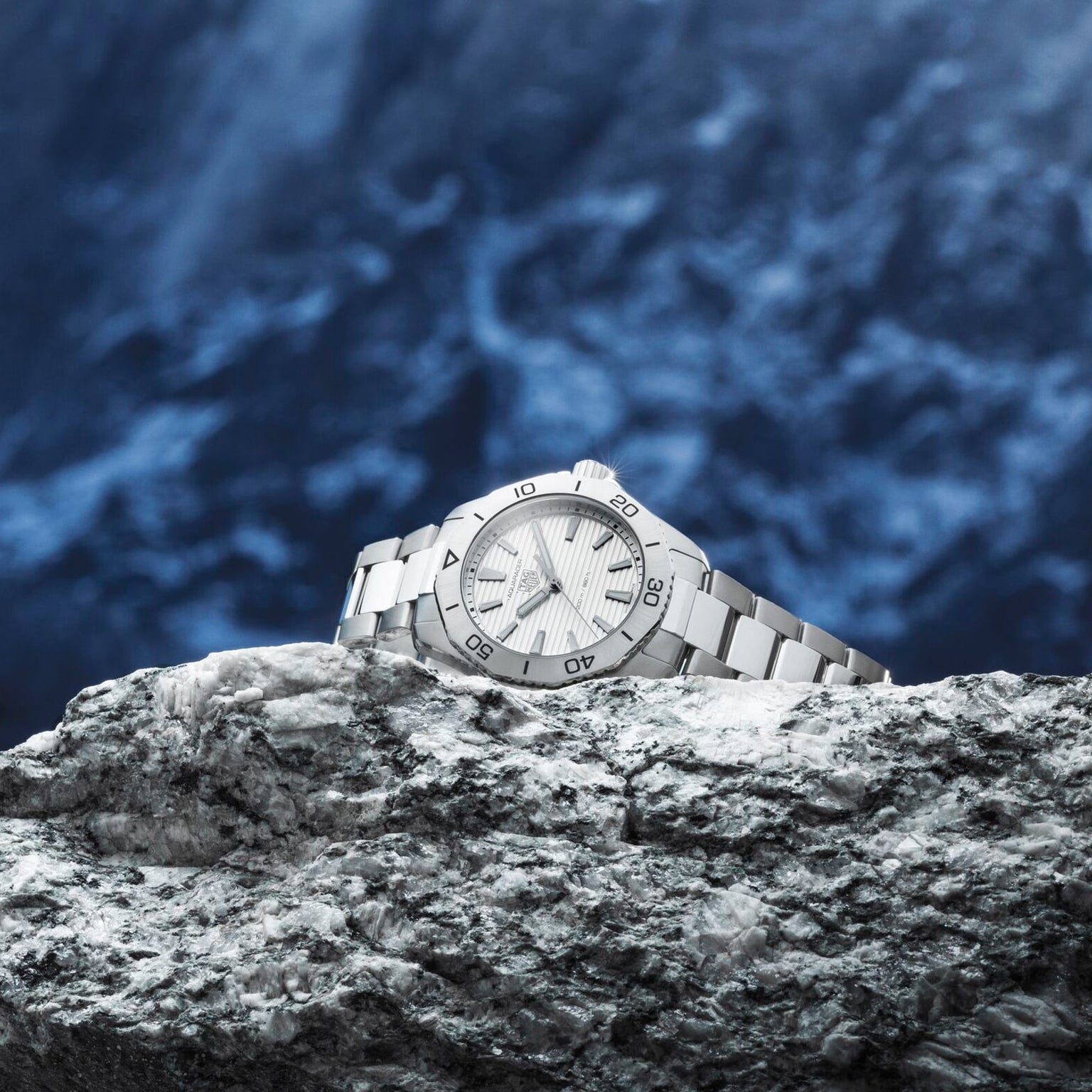 The TAG Heuer Aquaracer Professional 200 wristwatch with a stainless steel case is displayed on a textured gray rock, set against a blurred blue background.