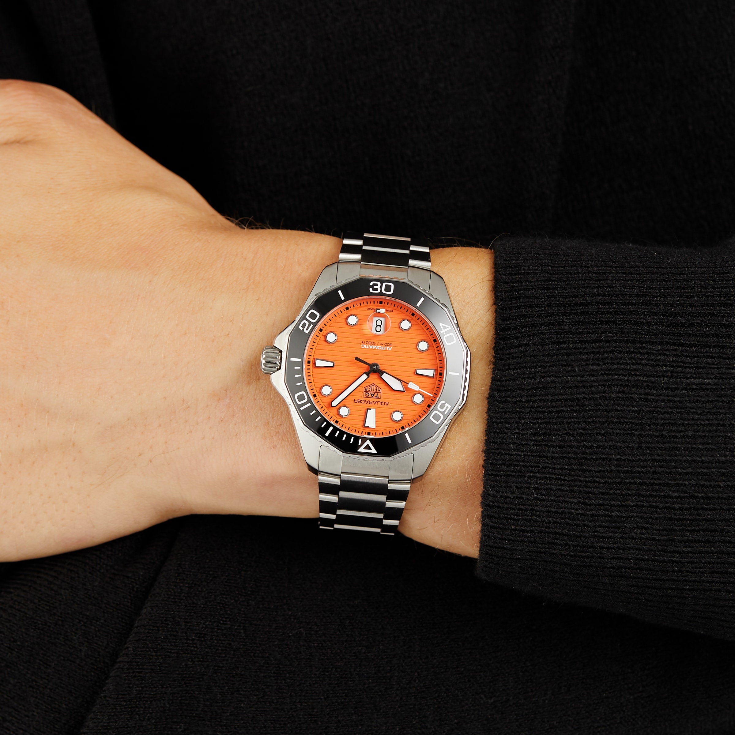 A wrist wearing the TAG Heuer Aquaracer Professional 300 Orange Diver, a silver and black watch with an orange face, with a black sleeve in the background.