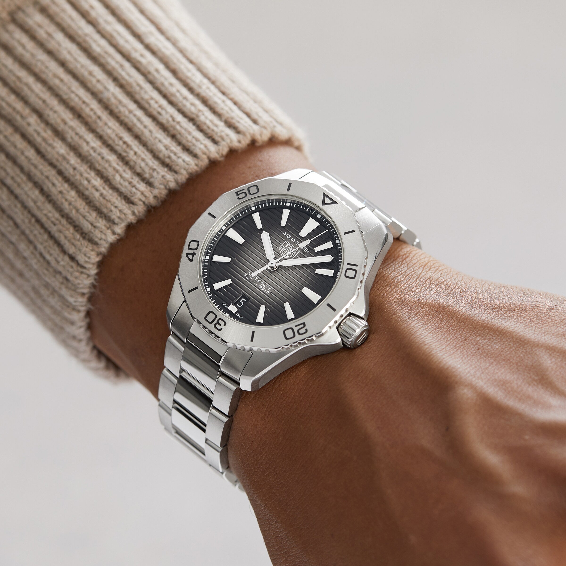 A TAG Heuer Aquaracer Professional 200 Date with a black face and white markers graces a person’s left wrist, complementing their beige ribbed sweater.