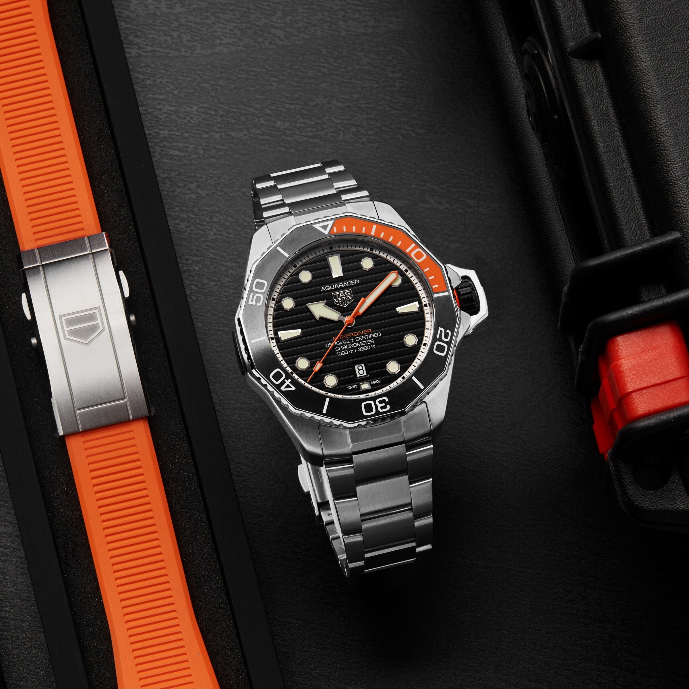 The TAG Heuer Aquaracer Professional 1000 Superdiver, a stainless steel dive watch with black and orange bezel, sits on a dark surface beside an orange strap and black case with orange accents, highlighting its impressive water resistance.