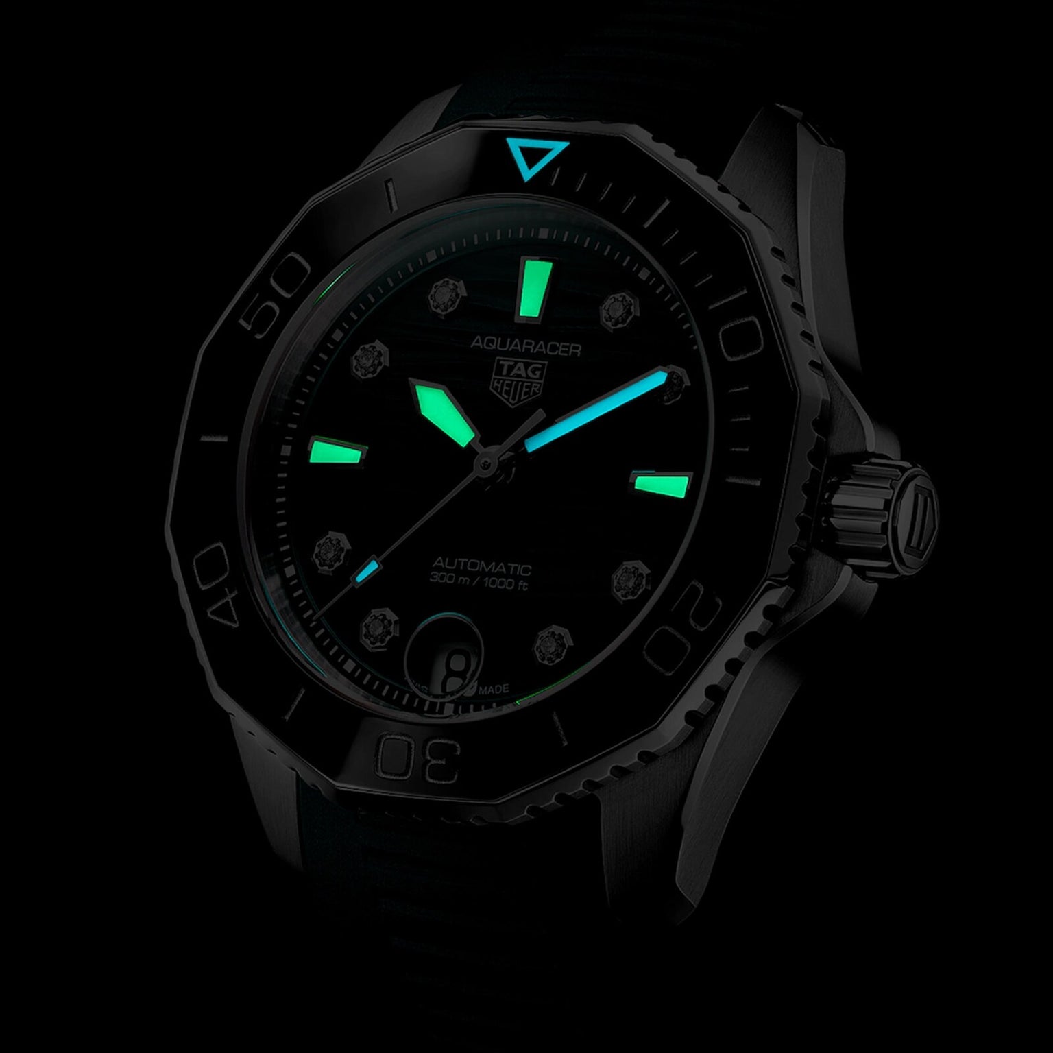 The TAG Heuer Aquaracer Professional 300 Date diving watch is displayed in a dark setting, showcasing its black design, glowing green hour markers and hands, luminous blue triangle at 12 o’clock, and striking blue lagoon dial accents.