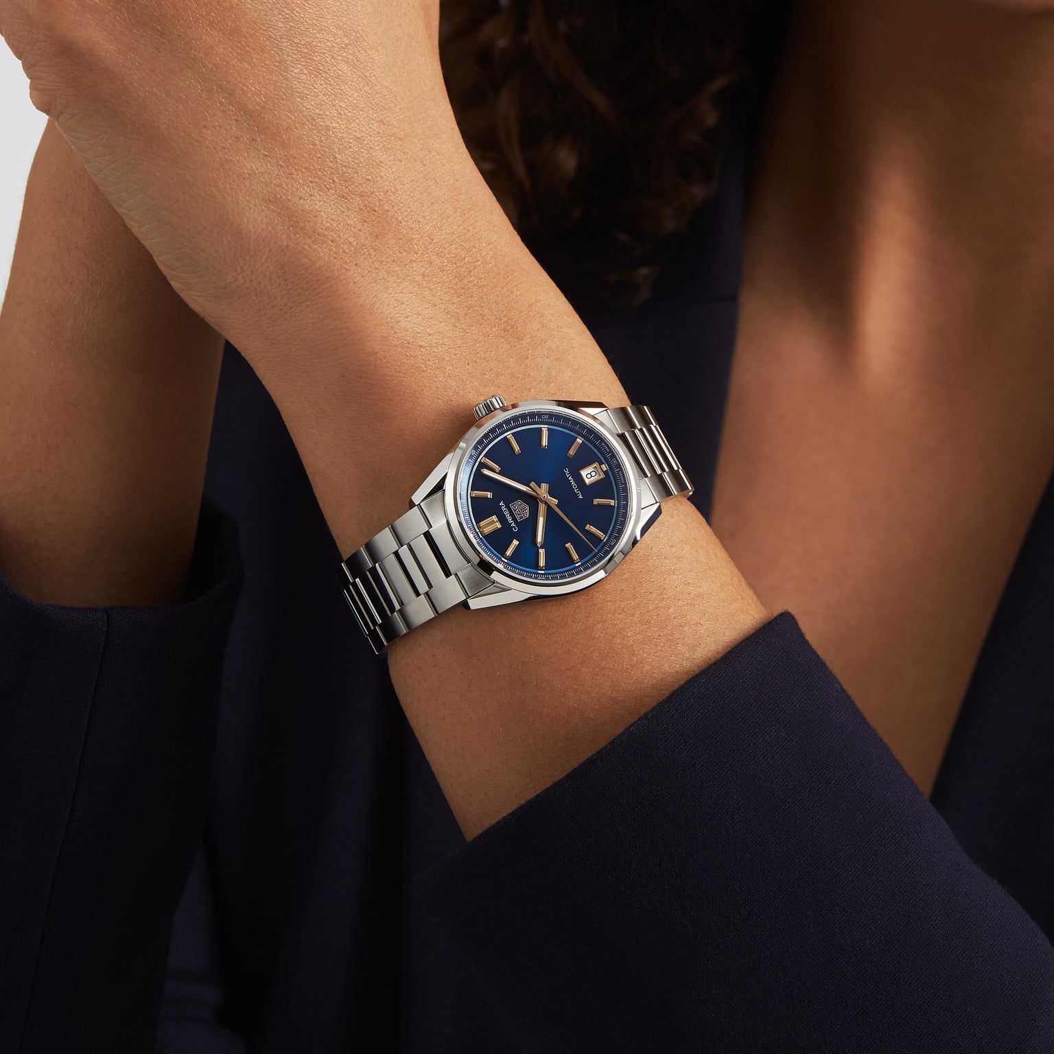 Dressed in a dark blazer, a person exudes elegance with one arm raised near their face, showcasing the TAG Heuer Carrera Date—a silver wristwatch with a blue dial and silver strap.