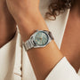 A person dressed in a cream blazer showcases the elegance of the TAG Heuer Carrera Date, a silver wristwatch featuring a light green face and date display.