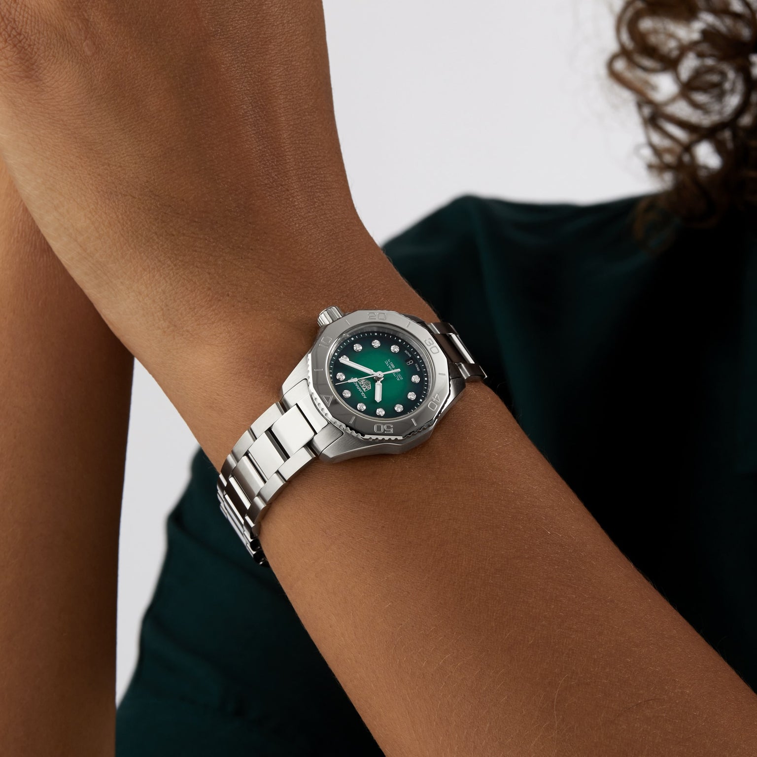A person wearing a TAG Heuer Aquaracer Professional 200 watch with a green face and silver band holds their hands together near their face against a neutral background.