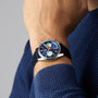 Wearing a blue textured sweater, a person showcases the TAG Heuer Carrera Chronograph Skipper, featuring a blue dial with colorful circular subdials, silver case, and black strap—design inspired by the Carrera Panamericana race.