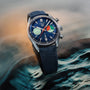 The TAG Heuer Carrera Chronograph Skipper, featuring a blue dial, multicolored subdials, and blue fabric strap, is displayed on a watery surface—a nod to the adventurous Carrera Panamericana race.