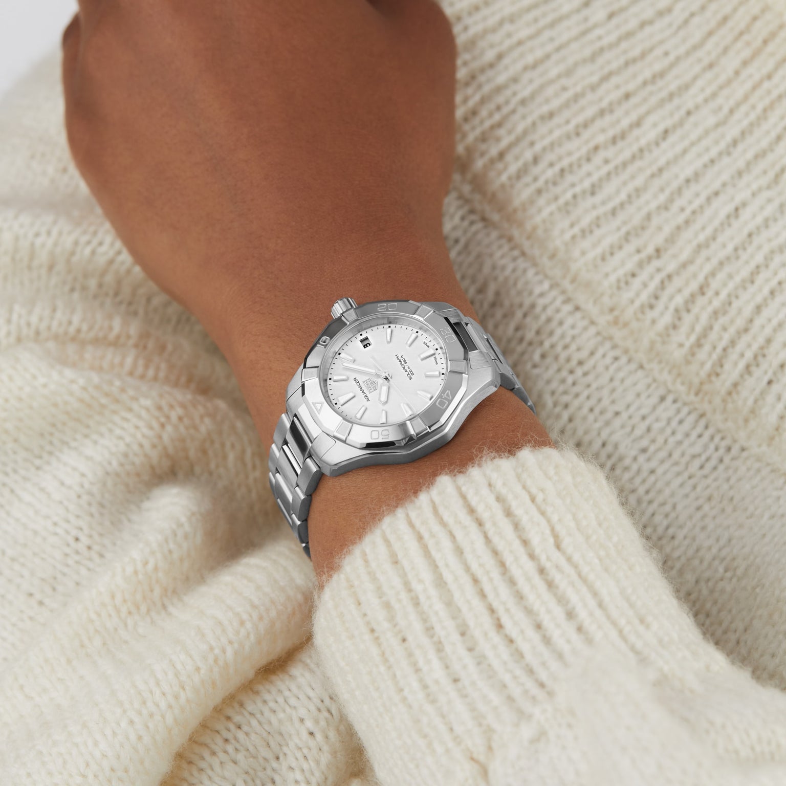 A TAG Heuer Aquaracer 200 Professional Solargraph silver wristwatch with a white face is worn on a wrist over a cream-colored knit sweater.