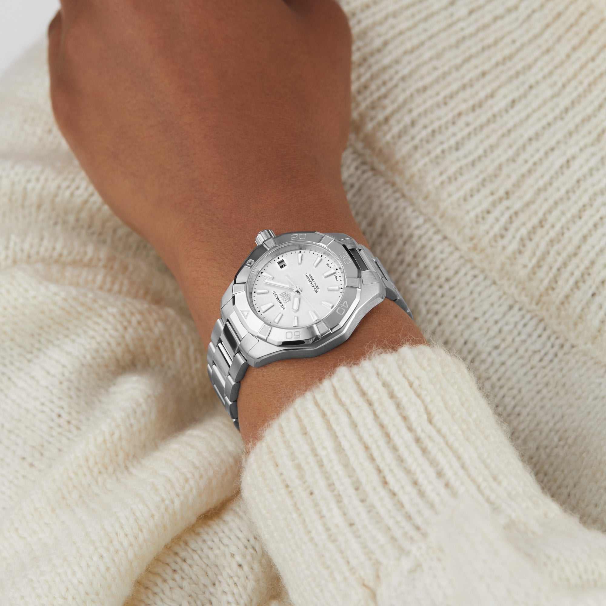A TAG Heuer Aquaracer 200 Professional Solargraph silver wristwatch with a white face is worn on a wrist over a cream-colored knit sweater.